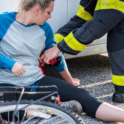 A woman sits on the ground beside a bicycle, receiving medical assistance from a firefighter.