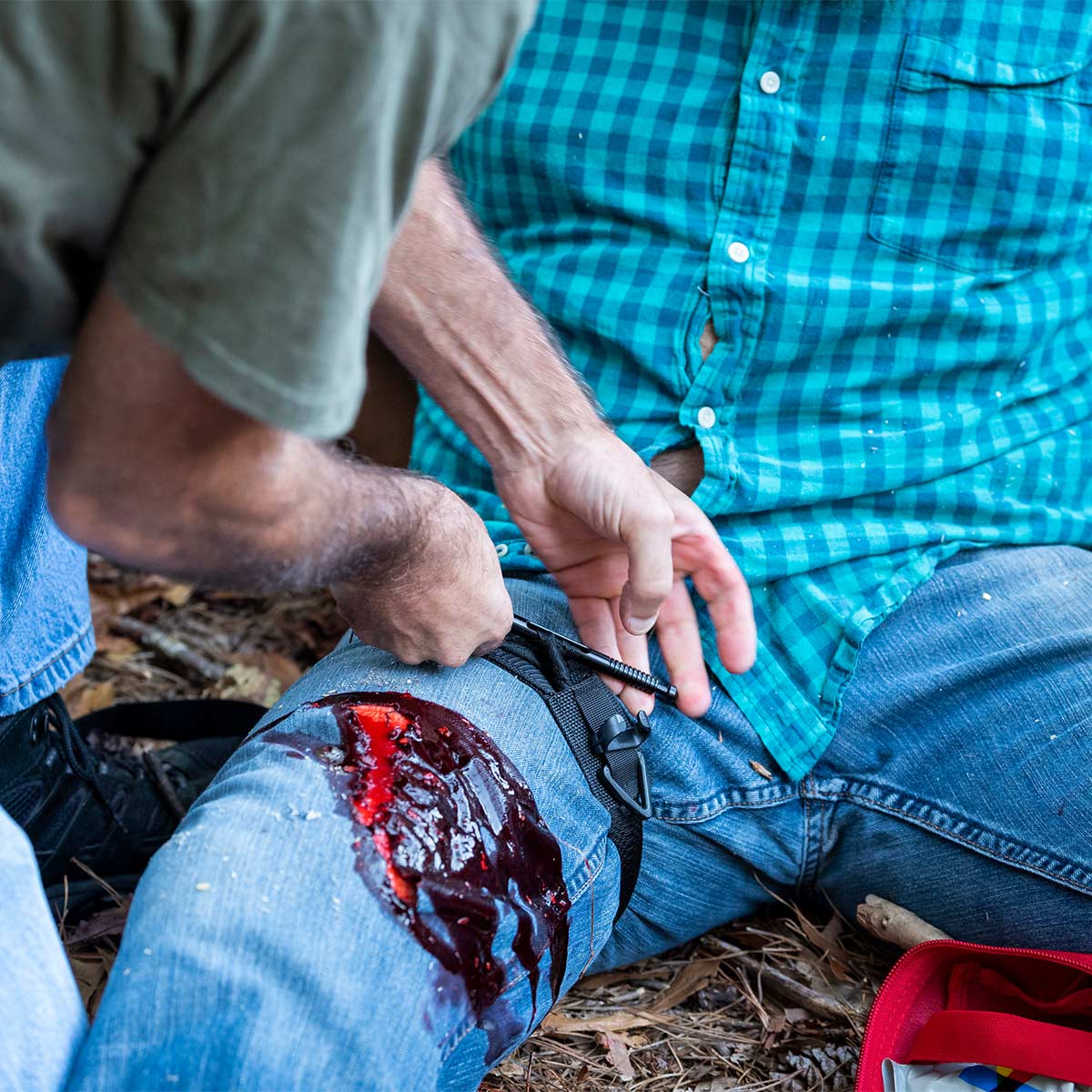 A person in a green shirt applies a tourniquet to a bleeding leg wound of someone in a blue plaid shirt.