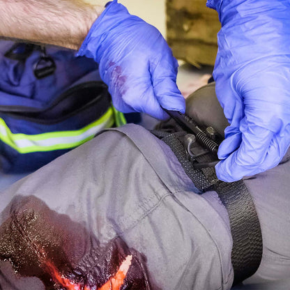 A police officer with a blue uniform applies a black tourniquet to their bloody arm, indicating an injury.