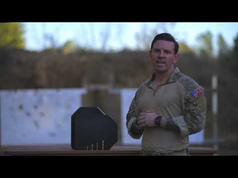 A person in military attire stands in front of a shooting range, next to a black protective target