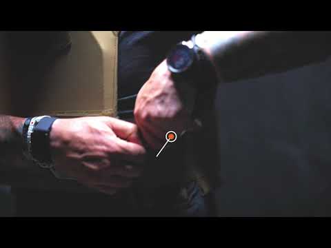 Two hands adjust a strap on a tan tactical vest in a dimly lit setting. The person wears a black shirt and wristwatch, evoking a focused and tactical mood.