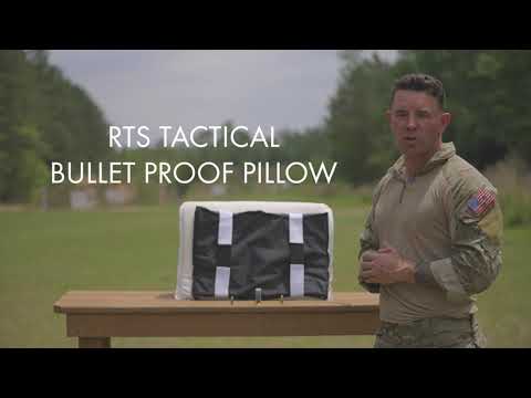 Man in tactical uniform stands beside a bulletproof pillow on a wooden table outdoors.