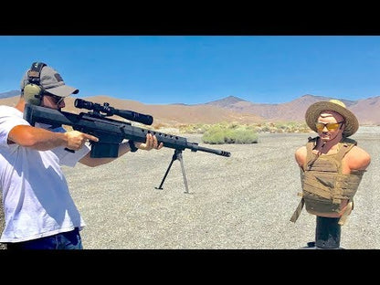 Man aiming a large rifle at a sand-filled mannequin wearing a cowboy hat and vest in a desert setting.