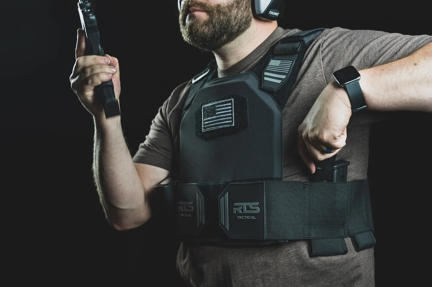 Man wearing tactical gear and earmuffs, holding a firearm magazine, with an RTS Tactical vest displaying a monochrome flag patch.