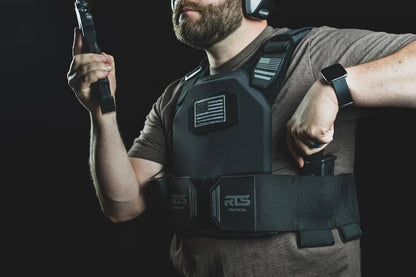Man wearing tactical gear and earmuffs, holding a firearm magazine, with an RTS Tactical vest displaying a monochrome flag patch.