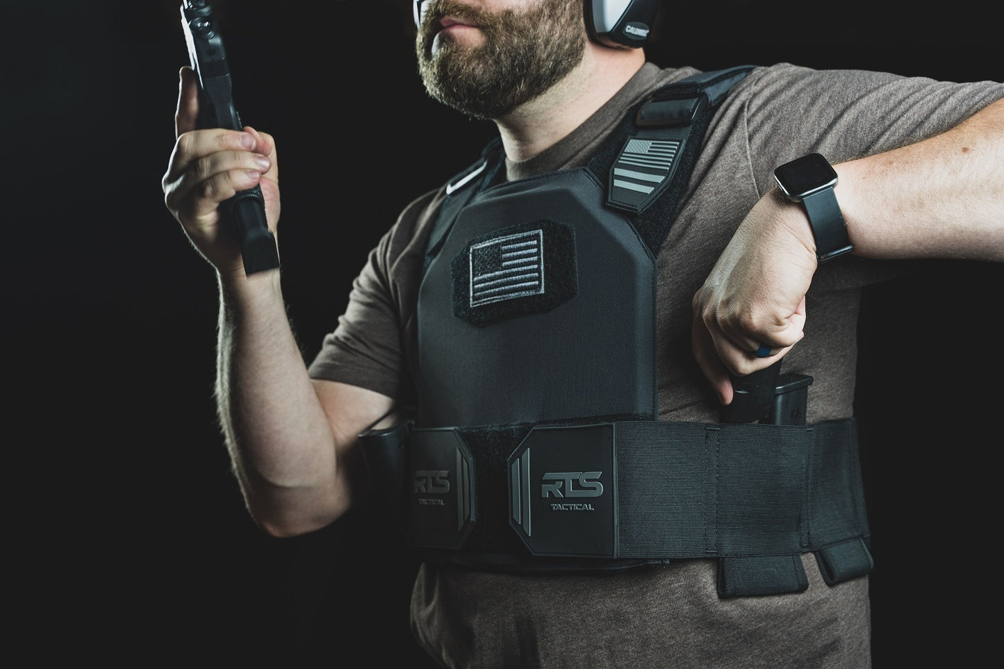 Man wearing tactical gear and earmuffs, holding a firearm magazine, with an RTS Tactical vest displaying a monochrome flag patch.