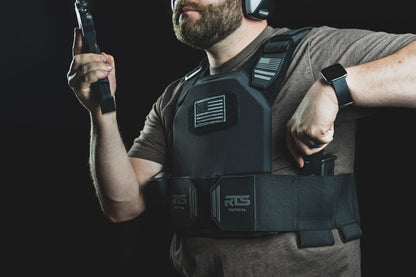 Man wearing tactical gear and earmuffs, holding a firearm magazine, with an RTS Tactical vest displaying a monochrome flag patch.